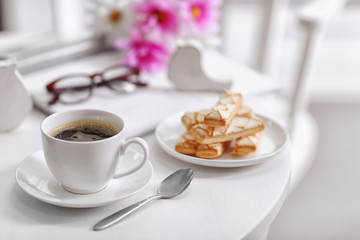 White cup of the coffee and fresh flowers on wooden table in a light room..