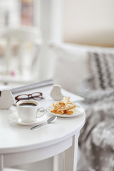 White cup of the coffee and sweets on wooden table  in a light room..