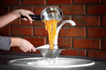Female hands pouring water from boiled pasta over sink in the kitchen - obrazy, fototapety, plakaty