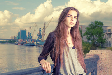 Lonely girl waiting for your. Wearing light gray shirt, black jacket, an young american woman standing by metal fence on pier in New York, frowned, port cranes on background. Instagram effect.