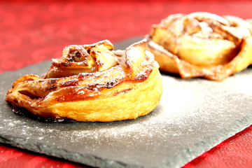 Two little roses of puff pastry with apples, cinnamon and powdered sugar on a slate dish. Red background.