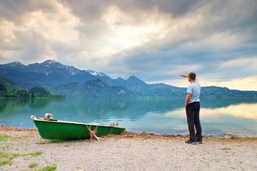Adult man in blue shirt walk at old fishing paddle boat at mountains lake coast.