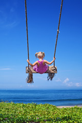 Little baby girl fly high with fun on rope swing on blue sky background on sea beach with waves and...