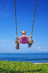 Little baby girl fly high with fun on rope swing on blue sky background on sea beach with waves and surf in tropical island. Travel lifestyle, people activity on summer family vacation with child.