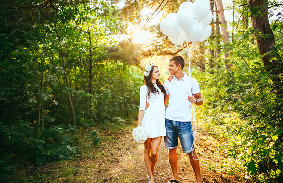 Young happy couple walking in the park. Man holing white baloons balloons. Woman hold white flowers boquet  