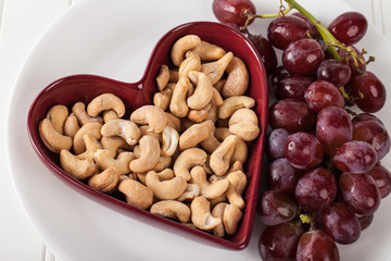 Cashews in a heart shaped bowl with red seedless grapes on white plate