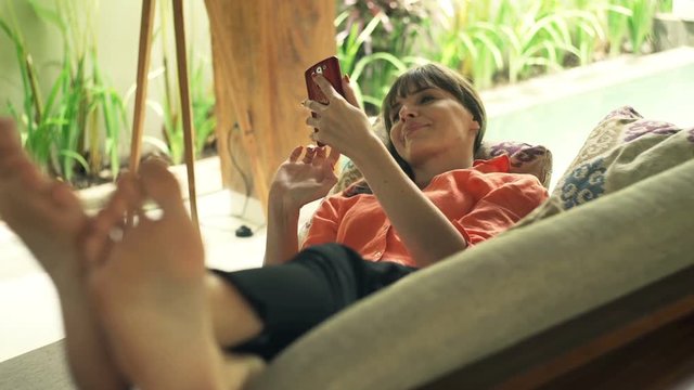Young, pretty woman using smartphone lying on sofa in outdoor villa
