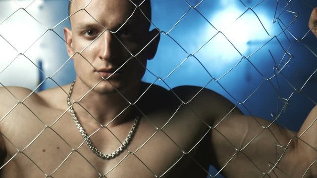 Sad bald topless man looking at camera while standing beyond wire fence in prison then looking down 