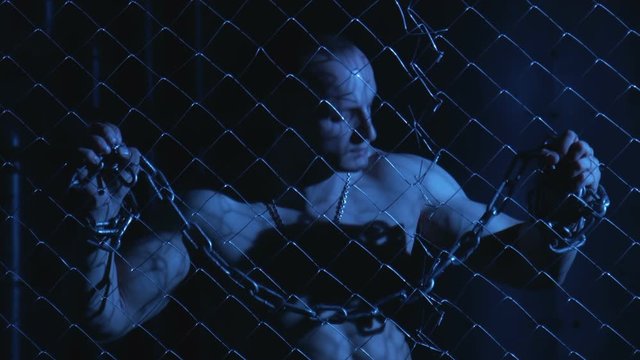 Topless sexy prisoner with metal chain in hands in moonlight holding fence while looking agressively at camera 
