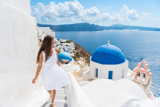 Fototapeta Santorini travel tourist woman on vacation in Oia walking on stairs. Person in white dress visiting the famous white village with the mediterranean sea and blue domes. Europe summer destination