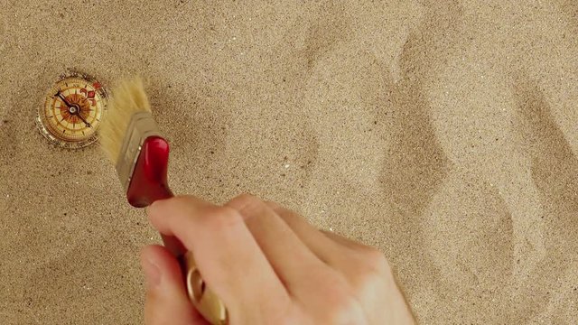 Discover vintage compass in desert sand, hand with brush, sweeping sandy dust.