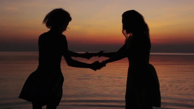 Silhouettes of two happy girls whirling at the beach on sunset. Slow motion