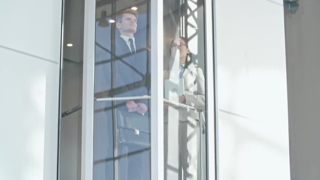 Low angle view of Caucasian man and Asian woman ascending in glass elevator inside business center. Suitable for business, office life, career growth, urban lifestyle visuals