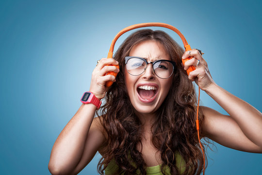 Angry crazy girl in headphones listening to music and screaming loud. Closeup portrait girl on blue background. Negative human emotion facial expression feeling