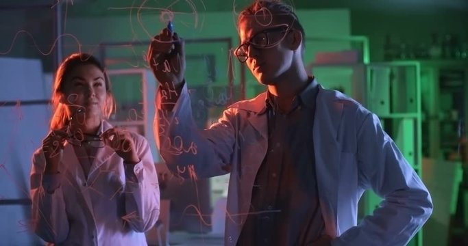 Two chemistry students working together in laboratory. Man drawing scheme on glass wall and explaining it to girl