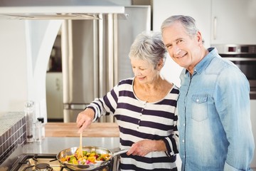 Portrait of senior man standing with wife cooking food