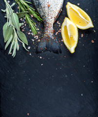 Tail of fresh raw Dorado or sea bream fish on black slate stone board with spices, herbs, lemon and...