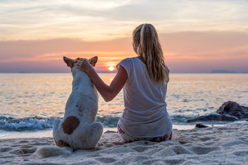 Fototapeta premium Young woman with dog sitting on the beach and watching the sunset