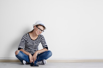 Stylish girl in hat and spectacles listening music with tablet pc at empty wall at home.  