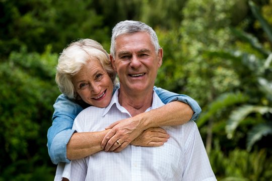 Senior Woman Embracing Husband From Behind Against Plants