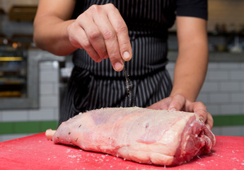 A fresh organic leg of lamb being seasoned with salt and pepper on a red chopping board.