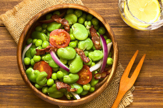 Frischer Salat aus Ackerbohnen, Erbsen, Kirschtomaten, rote Zwiebeln und Speck in Holzsch&uuml;ssel, fotografiert mit nat&uuml;rlichem Licht