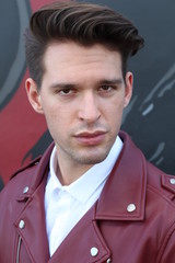 Bad boy concept. Portrait of brutal young man with short hair wearing red leather jacket, jeans and posing over urban background. Hand in pockets. Hipster style. Copy-space. Outdoor shot