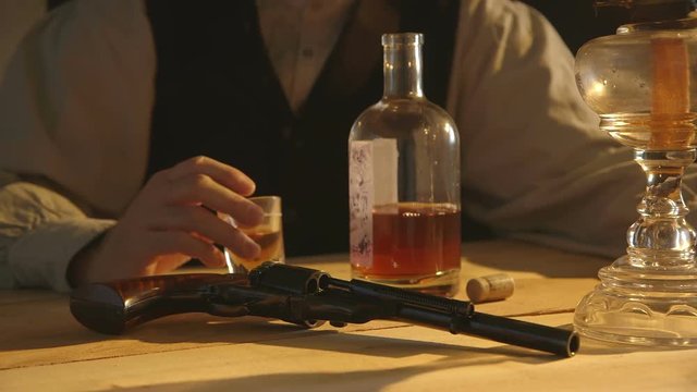 In a room lit by an oil lamp a cowboy from the American wild west era sitting at a rustic table builds his courage with whiskey.