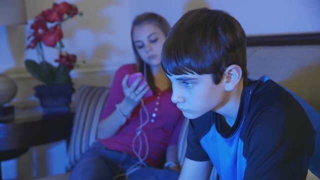 A young teen boy looks for something to watch on TV while his disinterested friend or sister sits using her smart phone.