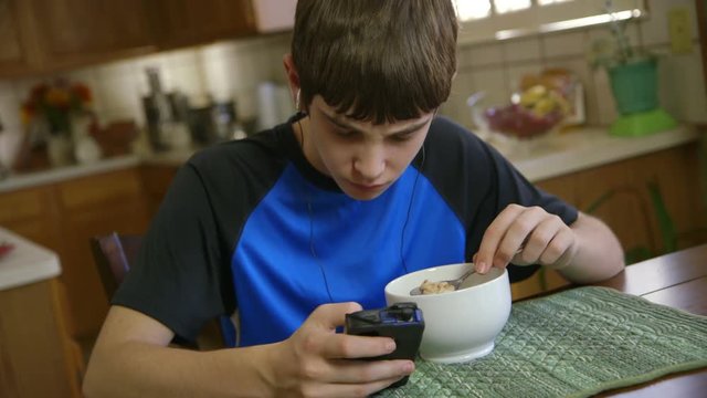 With his smart phone a teenage boy listens to music and reads text messages while he eats breakfast.