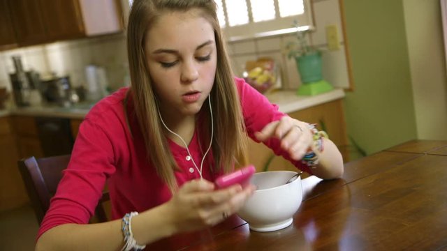 A pretty teenage girl eating at a kitchen looks up from her smart phone and smiles at the camera