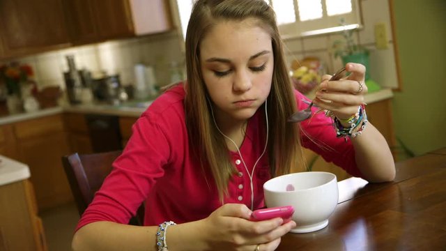 A pretty teenage girl eating at the kitchen listens to music and reads text messages on her smart phone.