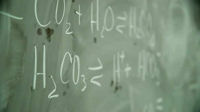 Scene focus changes from the end to the beginning of a chemical equation on a well used chalkboard.