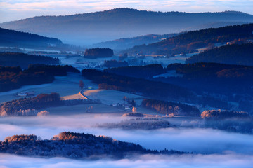 Fototapeta na wymiar Cold morning in Sumava National park, hills and villages in the fog and rime, misty view on czech landscape, blue winter scene, Tristolicnik, Czech Republic