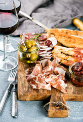 Meat appetizer selection. Salami, prosciutto, bread sticks, baguette, olives and sun-dried tomatoes, two glasses of red wine over grey concrete textured backdrop