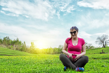 Naklejka na ściany i meble Young pregnant woman sitting on the grass at sunset and hugs your belly. 6 Month Pregnancy. The Concept Of Motherhood. Shows by means of hands sign heart. Beautiful view on blue sky background.