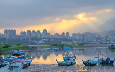 Morning landscape at dawn with sun beams shining through heavy clouds, stranded boats by riverside during a low tide & buildings in background ~ Beautiful sunrise scenery by Tamsui River Taipei Taiwan