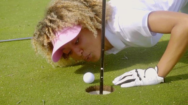 Young woman crouched down on the putting green blowing her golf ball into the hole as it sits a few inches from the lip of the cup.