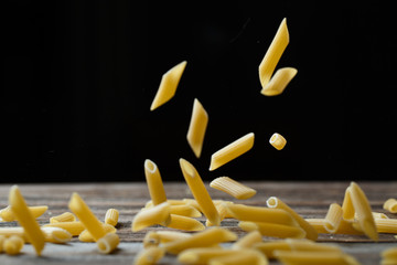 Falling penne pasta. Flying yellow raw macaroni over black background.