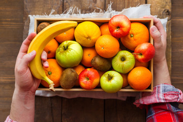Man in tartan plaid shirt holds a box full of fresh fruits and a banana.