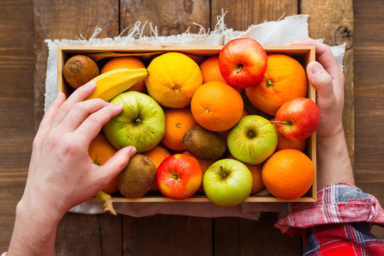 Man In Tartan Plaid Shirt Holds A Box Full Of Fresh Fruits. Fruit Harvest - Apples, Oranges, Banana, Kiwi.