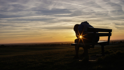 Sad unhappy silhouette man sitting with his head in his hands on a bench at sunset