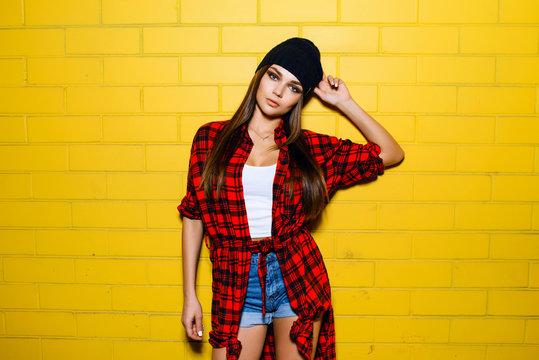 Beautiful young sexy hipster girl posing and smiling near urban yellow wall background in red plaid shirt, shorts, hat.