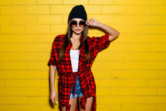 Beautiful young sexy girl posing and smiling near yellow wall background in sunglasses, red plaid shirt, shorts, hat.