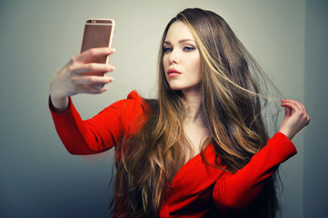Naklejka premium Beautiful woman in red dress with perfect cute face and blue eyes taking selfie with smartphone in studio.