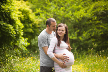 Happy couple waiting for baby. Man touches pregnant woman and hugs her belly outdoors in summer....