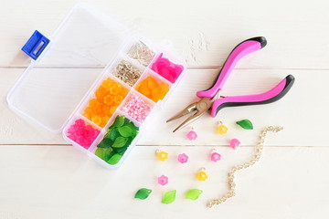 Organizer with beads, plastic flowers and accessories for handmade jewelry on white wooden background. Pliers, metal chain. How to make handmade bracelet. DIY concept