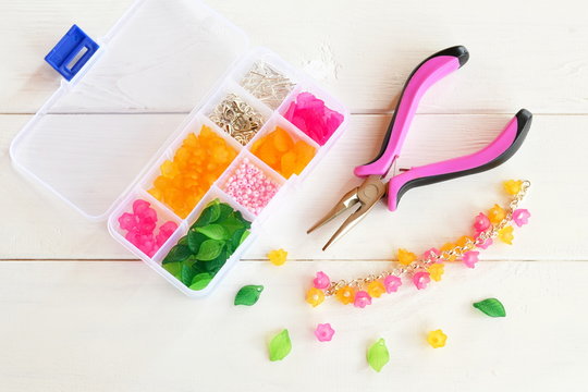 Organizer with beads, plastic flowers and accessories for handmade jewelry on white wooden background. Pliers. How to make handmade bracelet. DIY concept