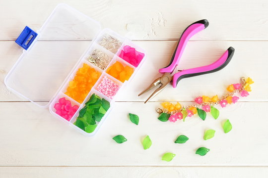 Organizer with beads, plastic flowers and accessories for handmade jewelry on white wooden background. Pliers. How to make handmade bracelet. DIY concept