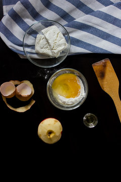 components of cooking cottage cheese cake (cottage cheese, flour, apple, egg, oil) on a black background